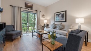 A living room with a sofa, armchairs, a coffee table, and a TV. There is a balcony with a glass door and curtains.