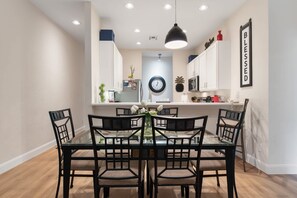 Un coin repas avec une table en verre, des chaises noires, un luminaire suspendu et une cuisine en arrière-plan.