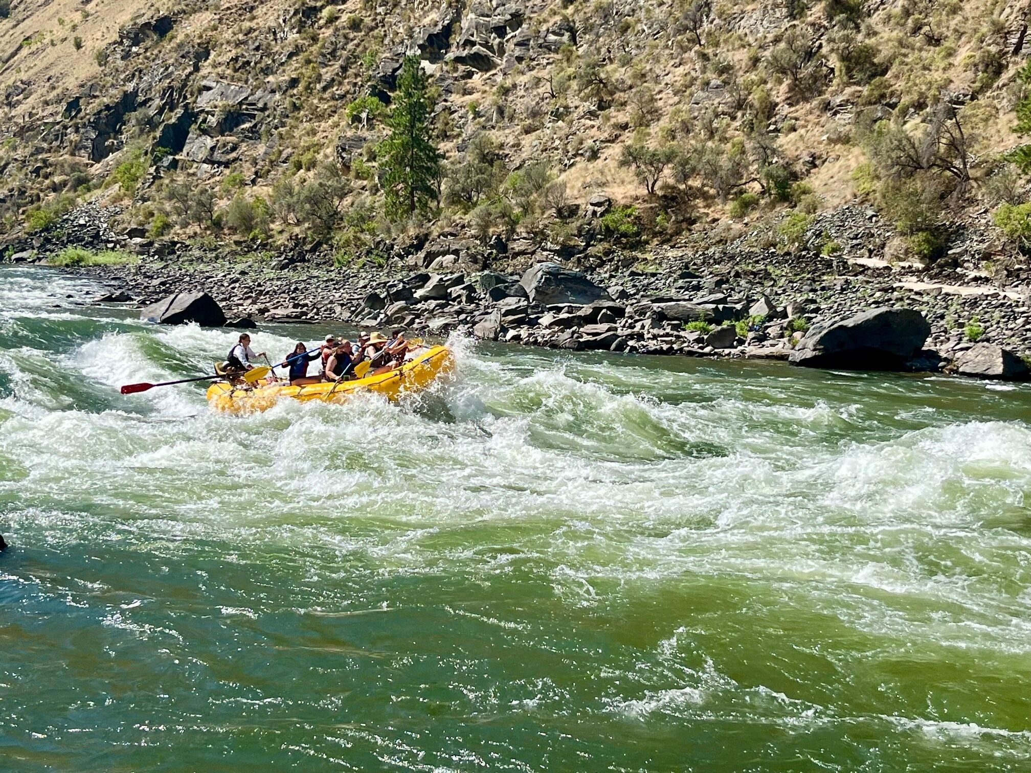 Een groep mensen in een gele raft die over een rivier vaart, omringd door rotsachtig terrein en schaarse begroeiing.