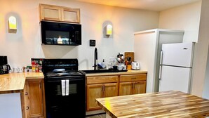 A compact kitchen with wooden cabinets, a black stove, a microwave, a white refrigerator, and a wooden dining table with two stools. - Steelhead Lodge (Lucile)