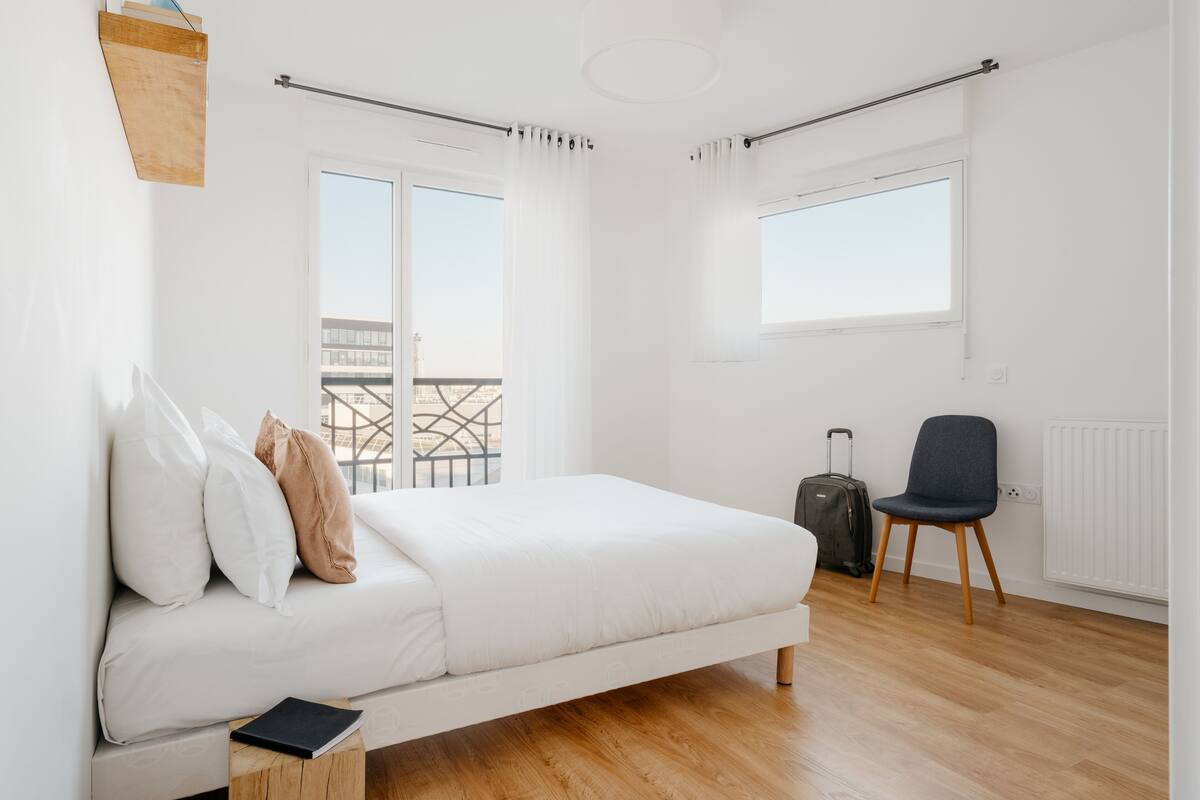 Een moderne slaapkamer met een bed, een stoel, een koffer en uitzicht op een gebouw.