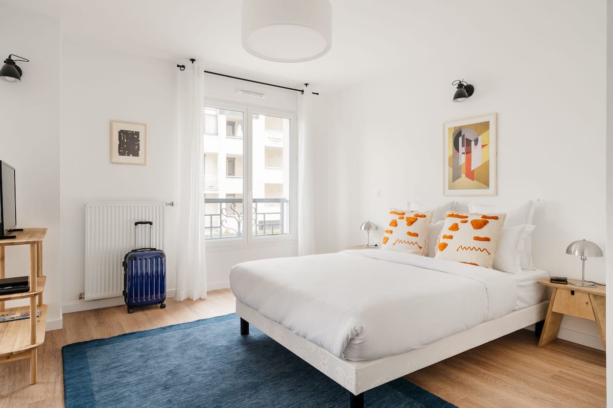 Een slaapkamer met een bed, een koffer, een radiator, een raam en kunstwerken aan de muur.