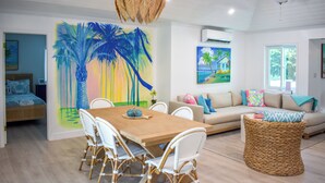 Uma sala de estar moderna com área de jantar, apresentando uma pintura grande de palmeiras, um sofá com almofadas coloridas e uma mesa de centro de vime.