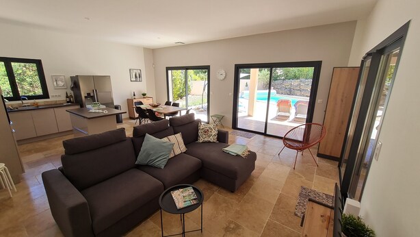 A modern living room with a dark brown sectional sofa, a small round table, and a dining area with a wooden table and chairs. - Recent villa with air conditioning and private pool on the heights of Barjac (Barjac)