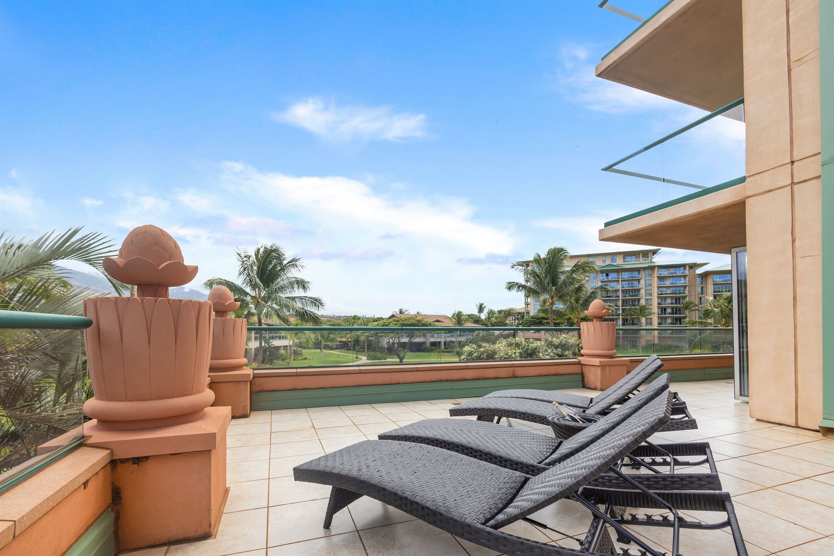 A balcony with lounge chairs, terracotta planters, and a view of a landscaped area with palm trees and buildings in the distance.