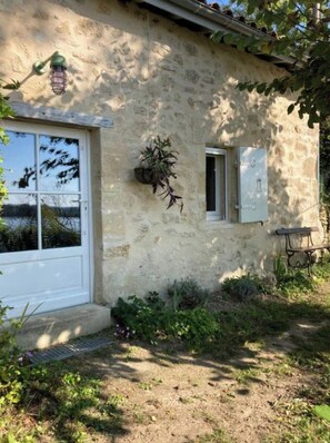 A rustic stone building with a white door and windows, a small bench, and a hanging plant.