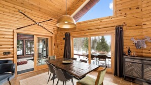 Interior de una cabaña de madera con una mesa de comedor, sillas, un sofá y un ventanal con vistas al exterior.