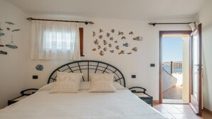 Een slaapkamer met een bed, twee kussens, een raam met gordijnen, een muur met visdecoraties, een deur naar een balkon en uitzicht op zee.