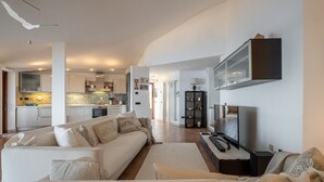 Een moderne woonkamer met een bank, een salontafel en een televisie. Achtergrond is een keuken met kasten en een fornuis.