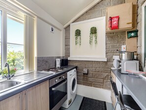 A compact kitchen with a built-in oven, microwave, and washing machine. There is a window with a view of greenery outside, and a shelf with a fire blanket and first aid kit. - Greystones Nawab Annex C (Flagg, Buxton)
