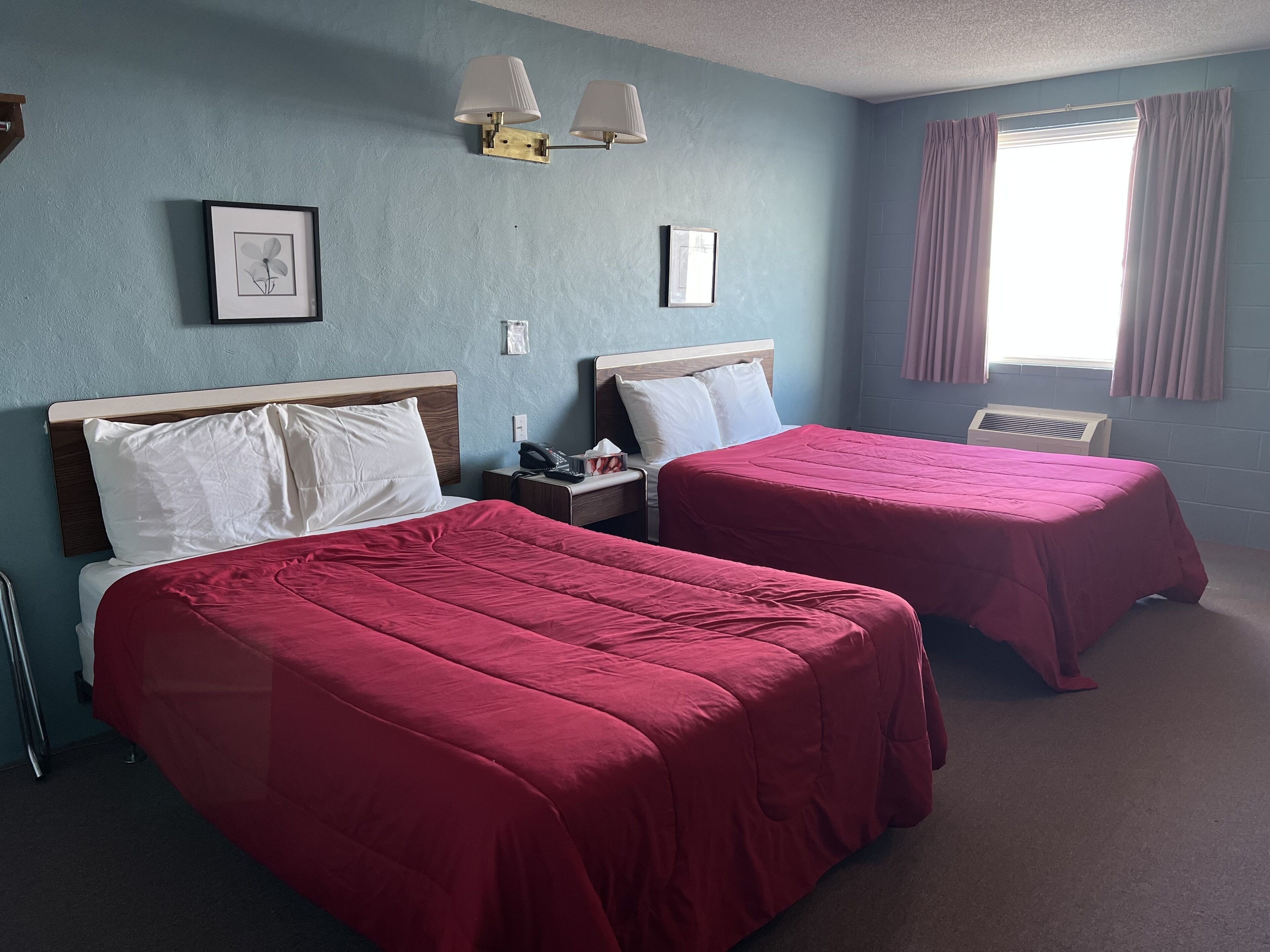 A hotel room with two beds, a nightstand, a wall-mounted lamp, and a window with curtains.