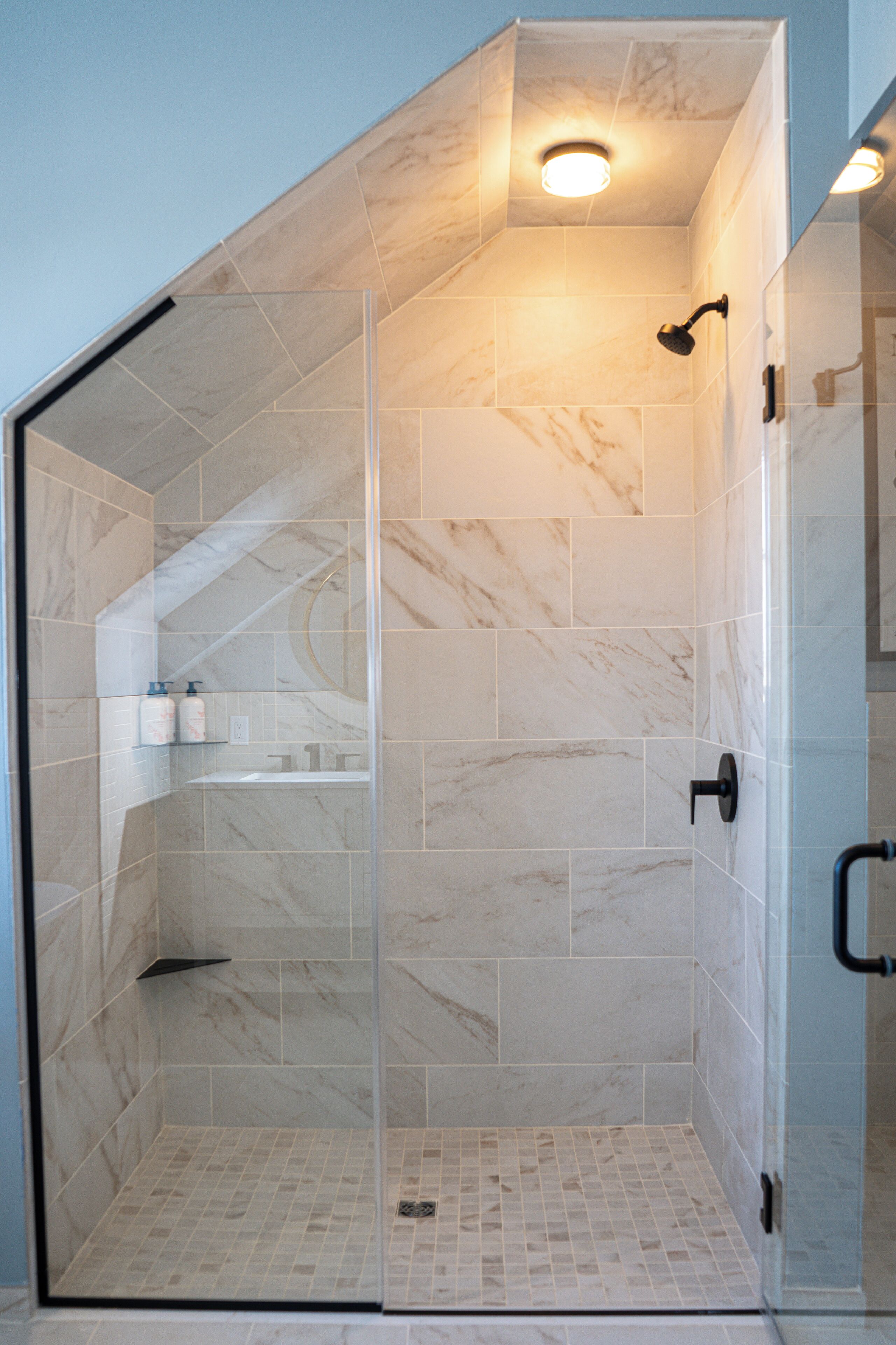 A glass-enclosed shower with a marble tile interior, a built-in shelf with toiletries, and a visible showerhead.