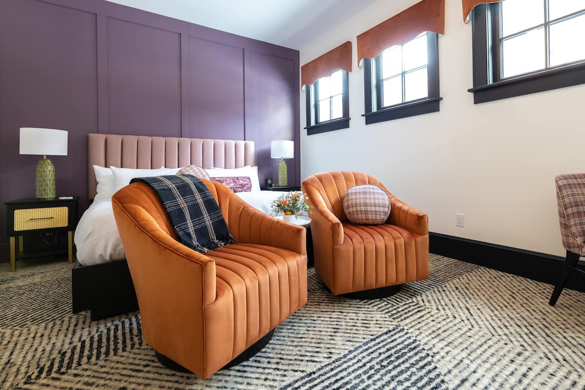 Une chambre d’hôtel avec un grand lit, deux fauteuils orange, un tapis à motifs et des murs violets.