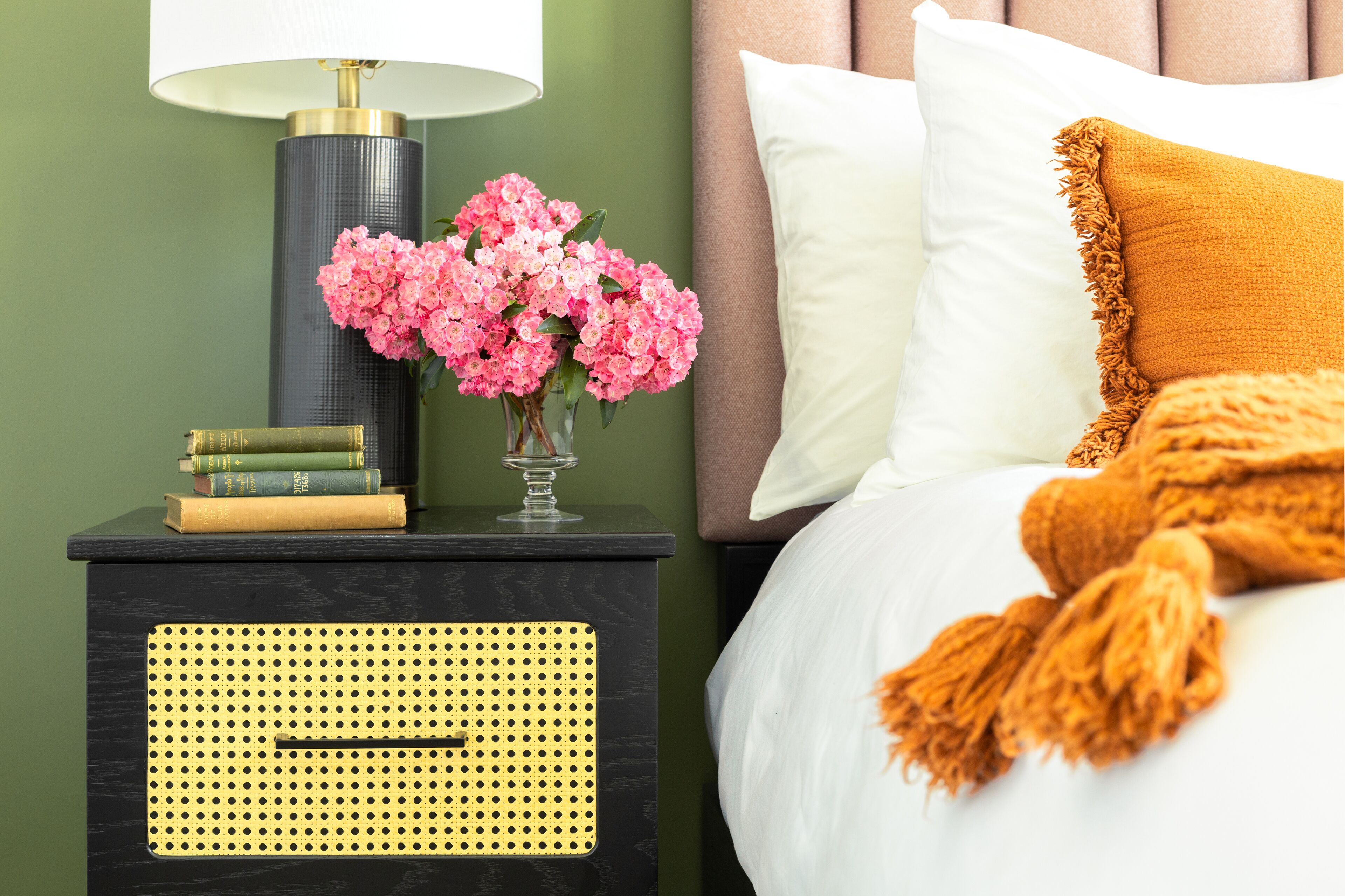 A neatly made bed with a white and mustard blanket, a bedside lamp, a vase of pink flowers, and a stack of books on a black nightstand.