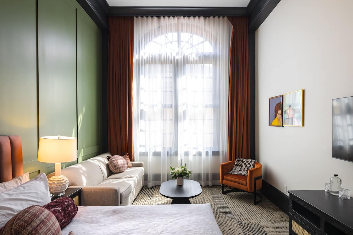 Une chambre d’hôtel avec un lit, un canapé, une chaise, une petite table sur laquelle se trouve un vase, une lampe et un téléviseur posé sur un meuble.