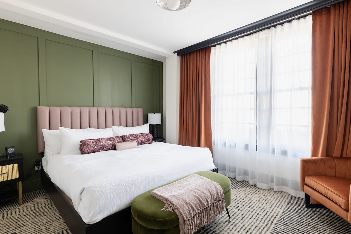 Une chambre d’hôtel bien aménagée, avec un grand lit, un pouf vert, un fauteuil marron et une fenêtre ornée de rideaux légers.