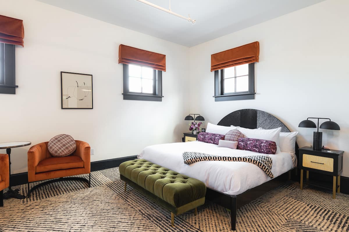 Une chambre à coucher comprenant un lit, un pouf, une chaise, une table de chevet et un tableau encadré accroché au mur.