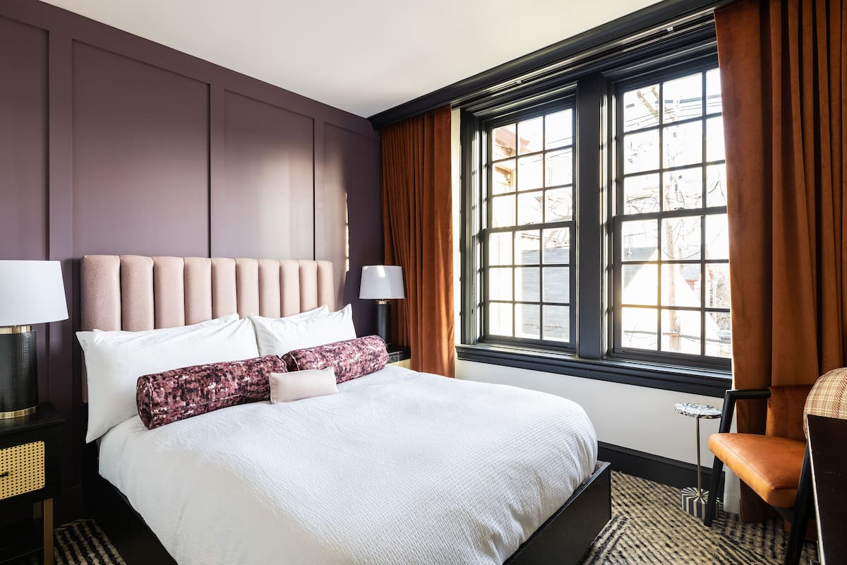 Une chambre d’hôtel avec un grand lit, une table de chevet avec une lampe, une chaise et une fenêtre avec des rideaux.