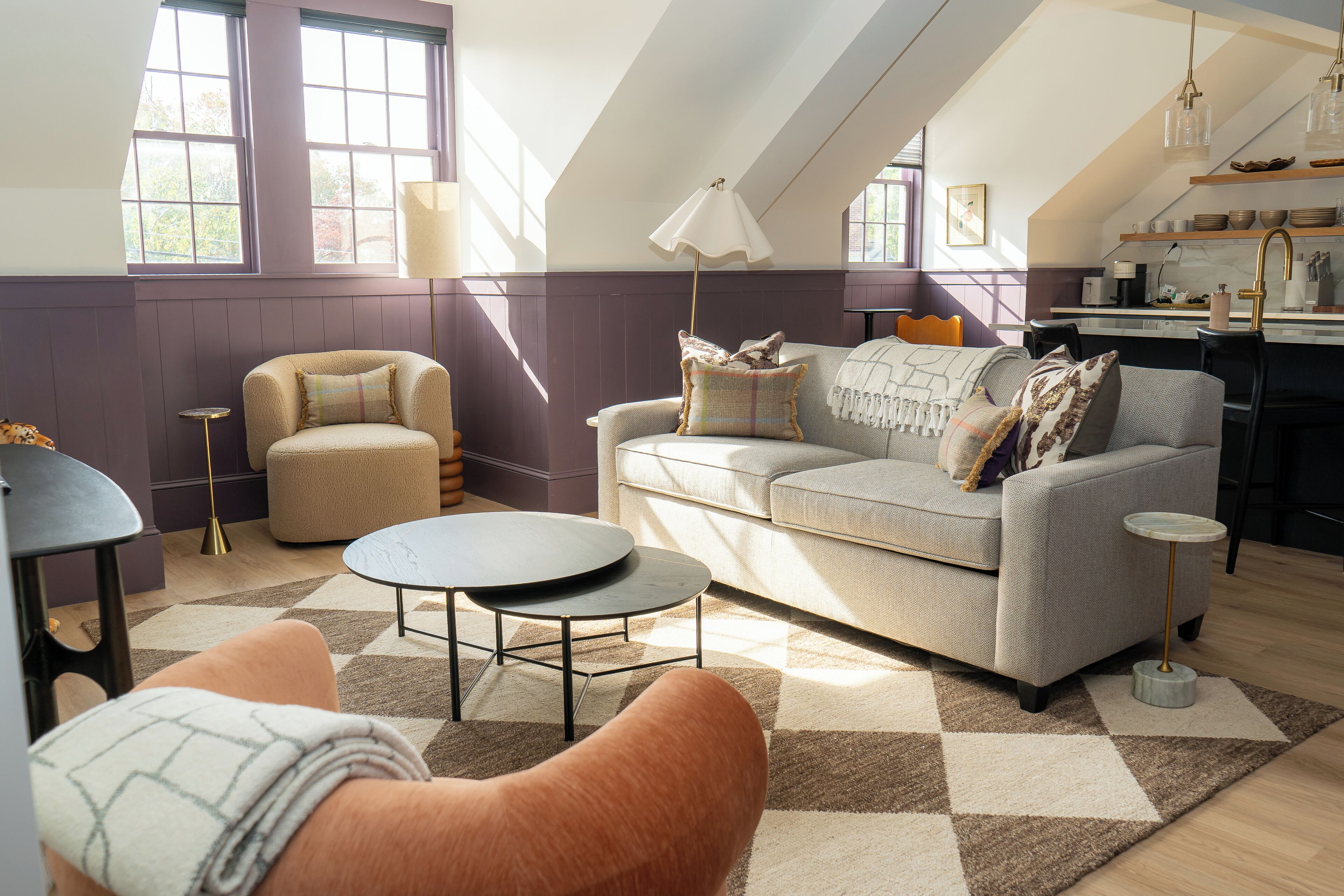 A modern living room with a sofa, armchair, and coffee table, a kitchen area with a sink and shelves, and large windows allowing natural light.