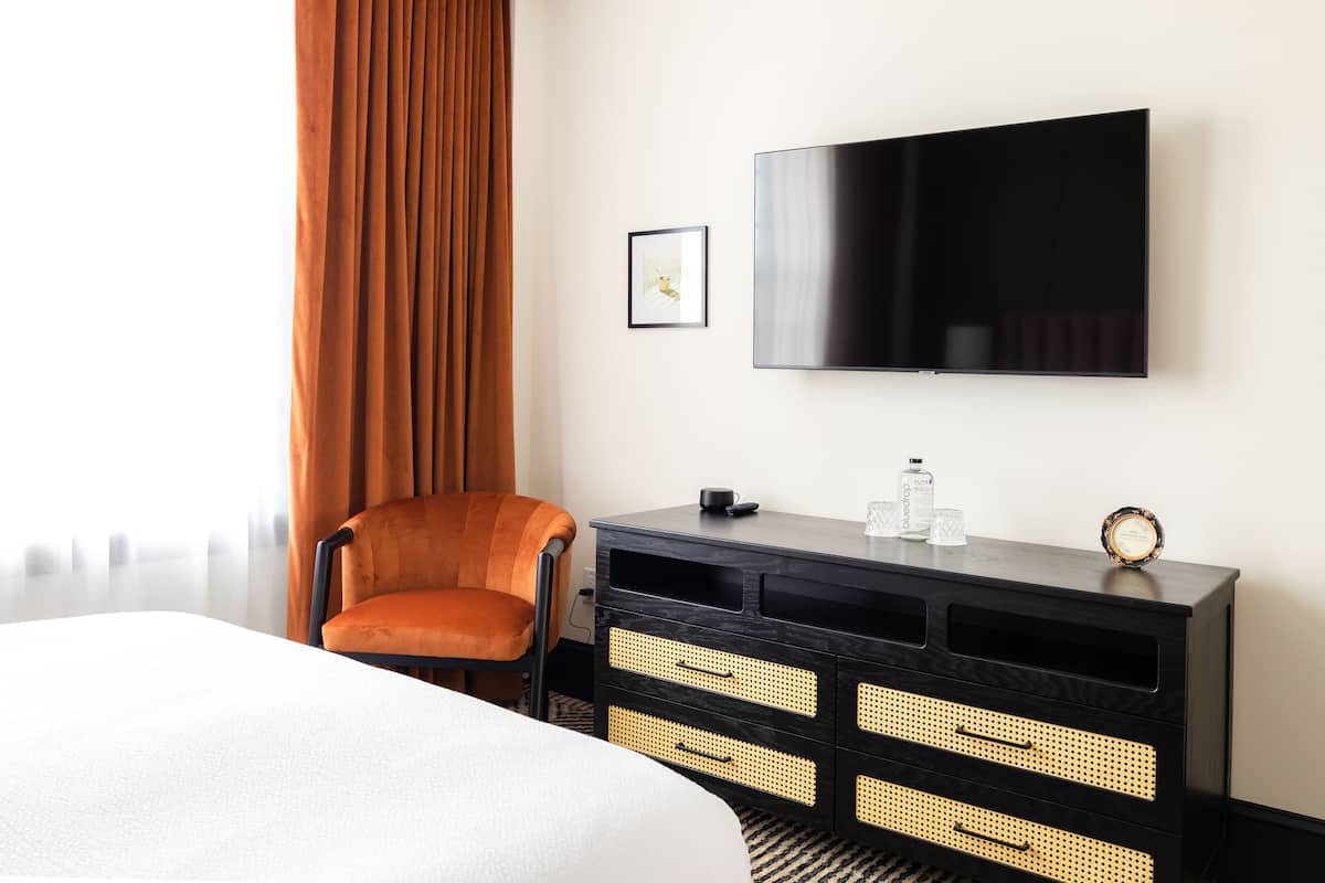 Une chambre d’hôtel avec un lit, une chaise, un téléviseur fixé au mur et une commode noire avec des tiroirs.