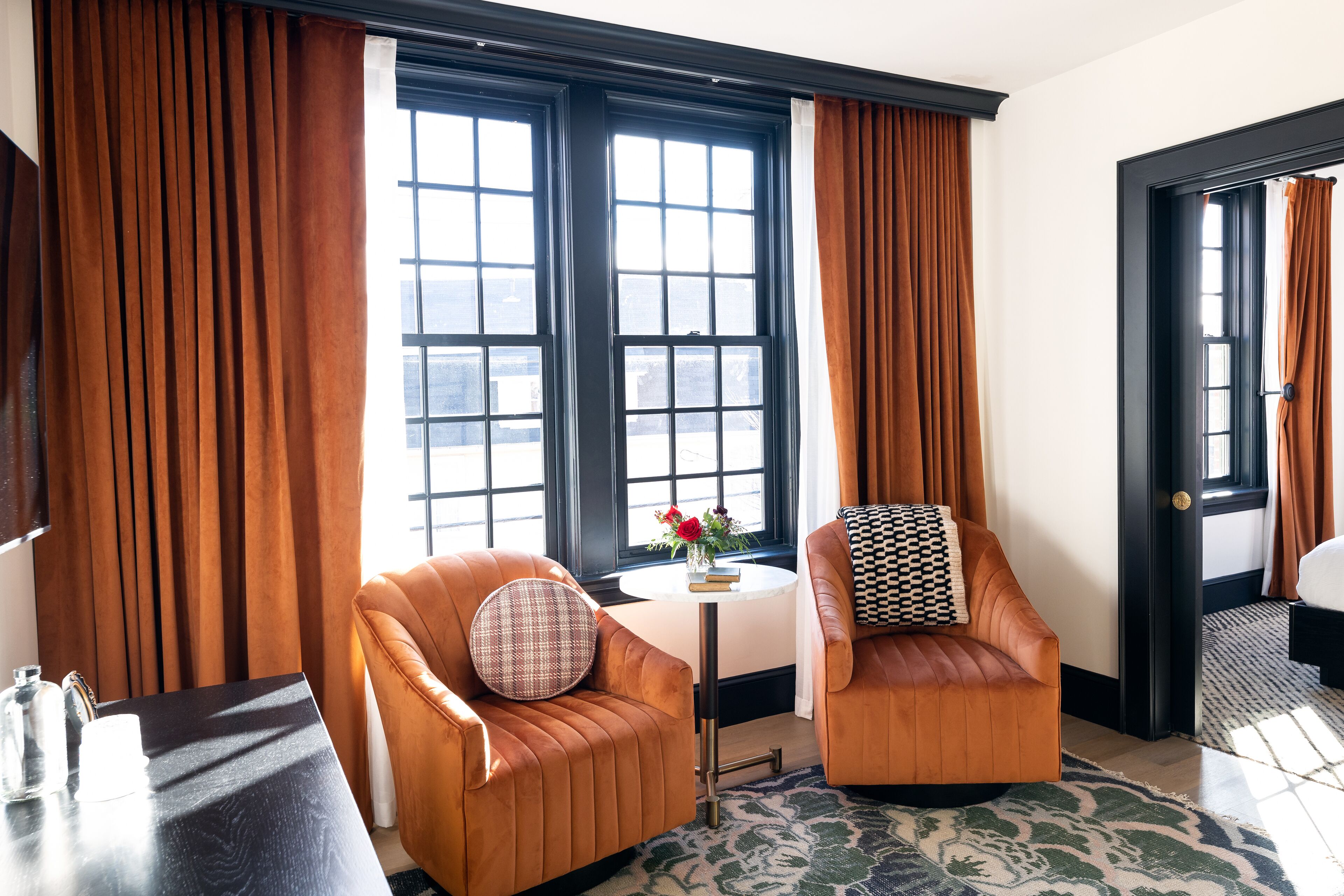 A hotel room with two leather armchairs, a small round table with a vase of flowers, and a view of the city through large windows with black frames.