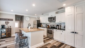 A modern kitchen with white cabinetry, a central island with stools, stainless steel appliances, and a coffee bar sign on the wall.