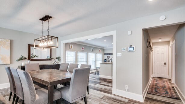A dining area with a wooden table and chairs, a chandelier, and a kitchen in the background.