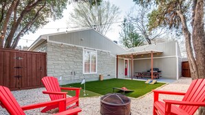 A backyard with a stone wall, red Adirondack chairs, a fire pit, and a patio area with string lights.
