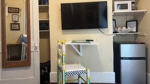Una habitación compacta con una televisión en la pared, un estante blanco pequeño con microondas y refrigerador.
