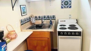 Una cocina compacta con estufa blanca, pileta y un mueble pequeño. Hay estantes sobre la pileta con vajilla y un azulejo decorativo en la pared.