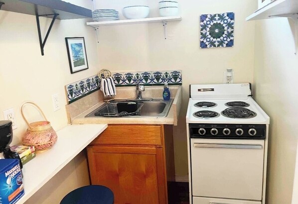 Una cocina compacta con estufa blanca, pileta y un mueble pequeño. Hay estantes sobre la pileta con vajilla y un azulejo decorativo en la pared.