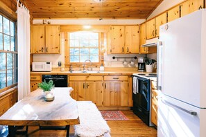Cocina de cabaña de madera con una mesa de madera, un refrigerador blanco, una estufa, un microondas y un lavamanos.