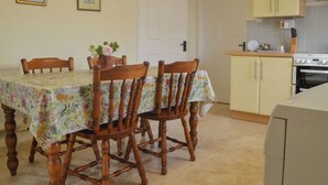 A dining area with a floral tablecloth, wooden chairs, and a vase with flowers. There is a kitchen with yellow cabinets and a stove.