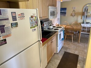 Una cocina equipada con refrigerador, estufa, microondas y gabinetes de madera. Se observan sillas y una mesa de comedor al fondo.