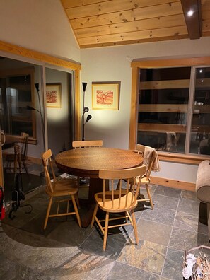 A dining area with a round wooden table and four chairs, large windows, and framed artwork on the walls.