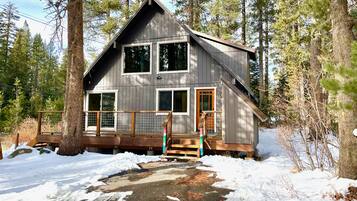 A two-story cabin with a wooden deck and a front porch, surrounded by trees and snow.