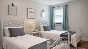 A bedroom with two white beds, a small bedside table with a lamp and books, a framed inspirational quote on the wall, and a window with curtains.