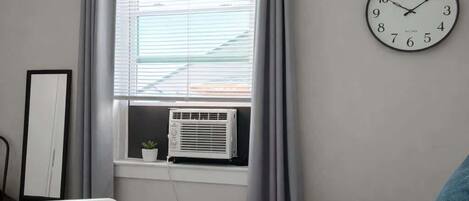 Un dormitorio con una cama, un reloj de pared, un ventanal con cortinas y un equipo de aire acondicionado.