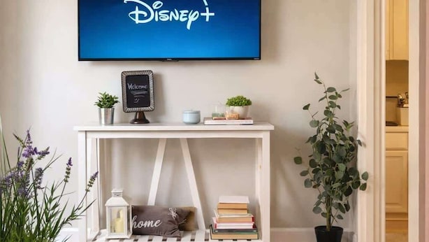 A living room with a flat-screen TV displaying the Disney+ logo, a white console table with decorative items, a potted plant, and a bookshelf.