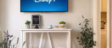 Ein Wohnzimmer mit einem Flachbildfernseher, der das Disney+-Logo zeigt, einem weißen Konsolentisch mit dekorativen Gegenständen, einer Topfpflanze und einem Bücherregal.