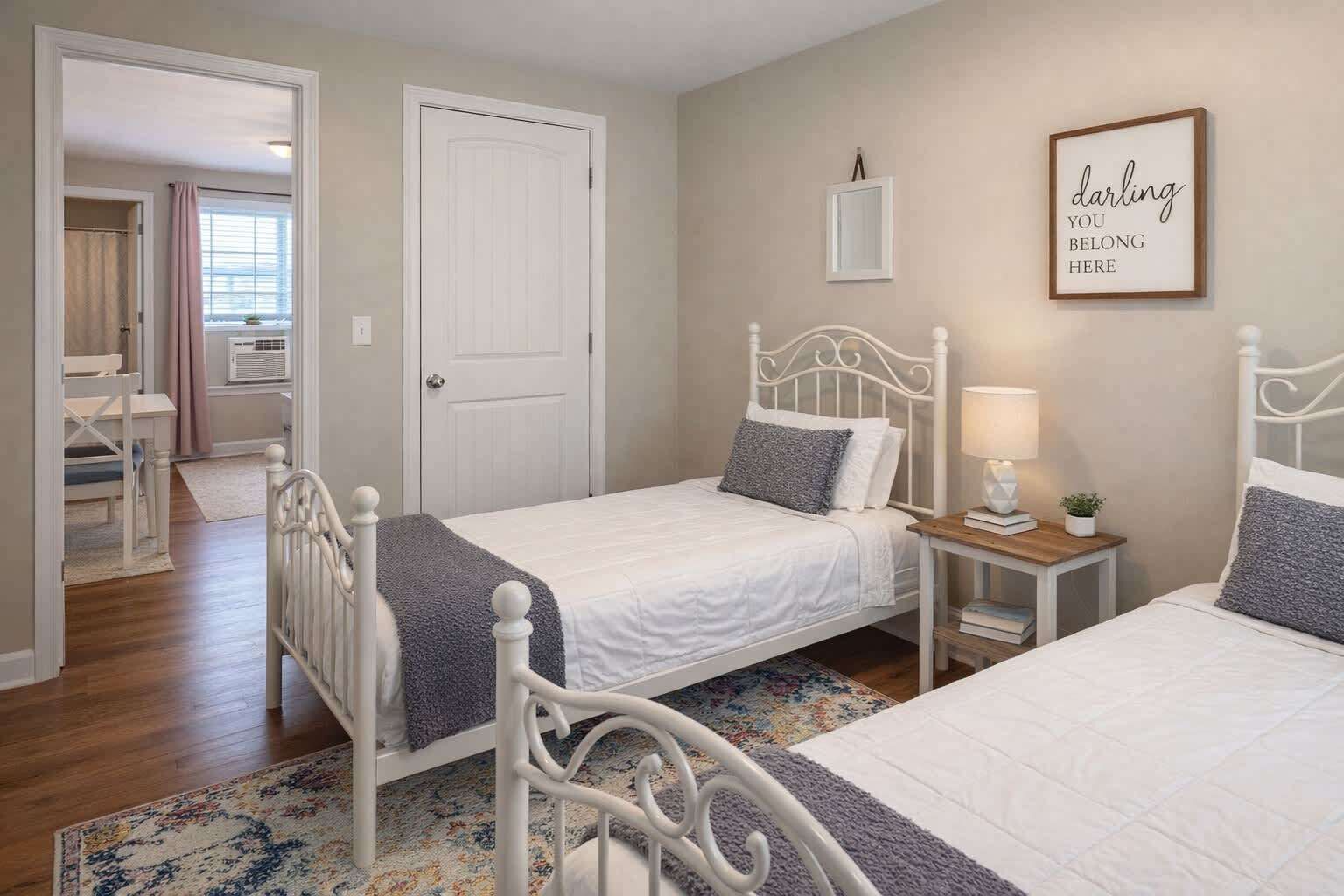A bedroom with two single beds, a bedside table with a lamp, a framed inspirational quote, and a view of a bathroom with a shower and a mirror.