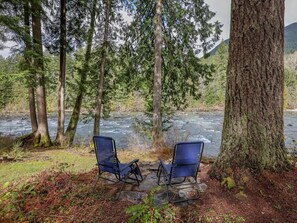 Twee blauwe stoelen staan op een stenen rand, met uitzicht op een rivier, omringd door dicht bos en hoge bomen.