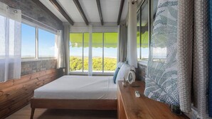 A bedroom with a wooden bed, a wooden nightstand, a large window with a view, and a patterned curtain. - Villa Priva • Meilly Estate (Saint-Louis)