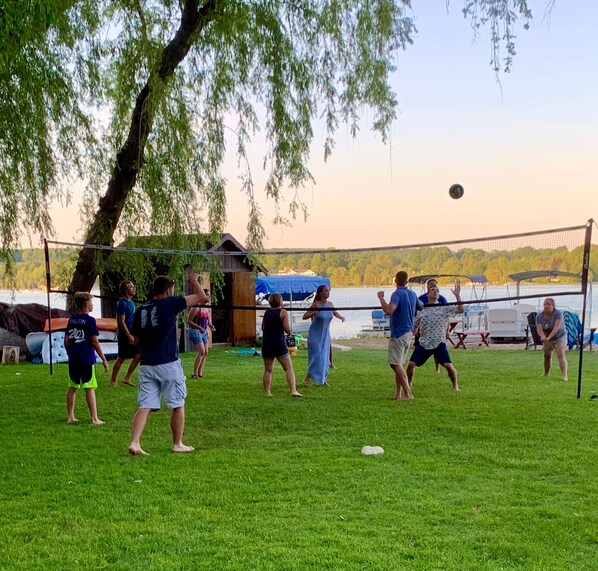 Un grupo de personas jugando voleibol en un campo cubierto de césped, con una red, árboles y un cuerpo de agua al fondo.