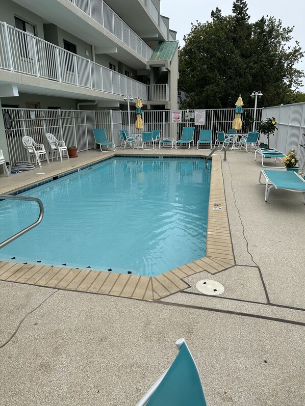 Une piscine entourée de chaises longues et de parasols, le tout délimité par une clôture, avec un bâtiment visible à l’arrière-plan.