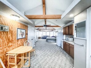 A compact, well-lit room with a wooden wall, a dining table with chairs, a kitchenette, and a bed with a blue bedspread.