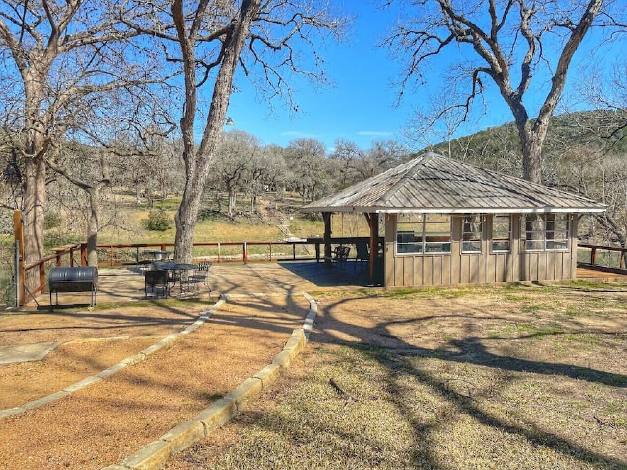 Un pabellón rústico de madera con un área de estar cubierta, rodeado por un jardín vallado con parrillas de barbacoa.