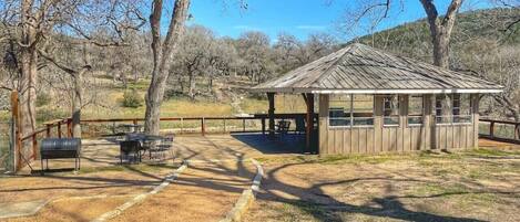 Un pabellón rústico de madera con un área de estar cubierta, rodeado por un jardín vallado con parrillas de barbacoa.