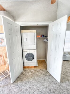 A compact laundry room with a washer and dryer, a small shelf with cleaning supplies, and a door leading to another room.