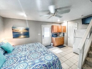 A bedroom with a bed, a ceiling fan, and a view of a kitchen with wooden cabinets and a refrigerator.